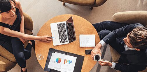 Two people sitting at a table with laptops and papers.